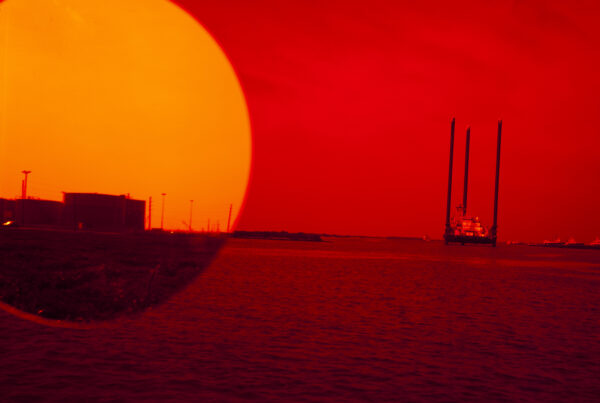 Photograph of a red sky and sea with boats at a distance. A large yellow circle on the top left resembles a sun setting over the ocean. Inside the circle, there is an image of a land mass with industrial buildings.