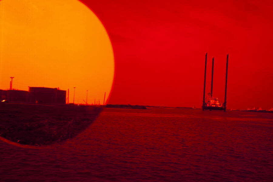 Photograph of a red sky and sea with boats at a distance. A large yellow circle on the top left resembles a sun setting over the ocean. Inside the circle, there is an image of a land mass with industrial buildings.
