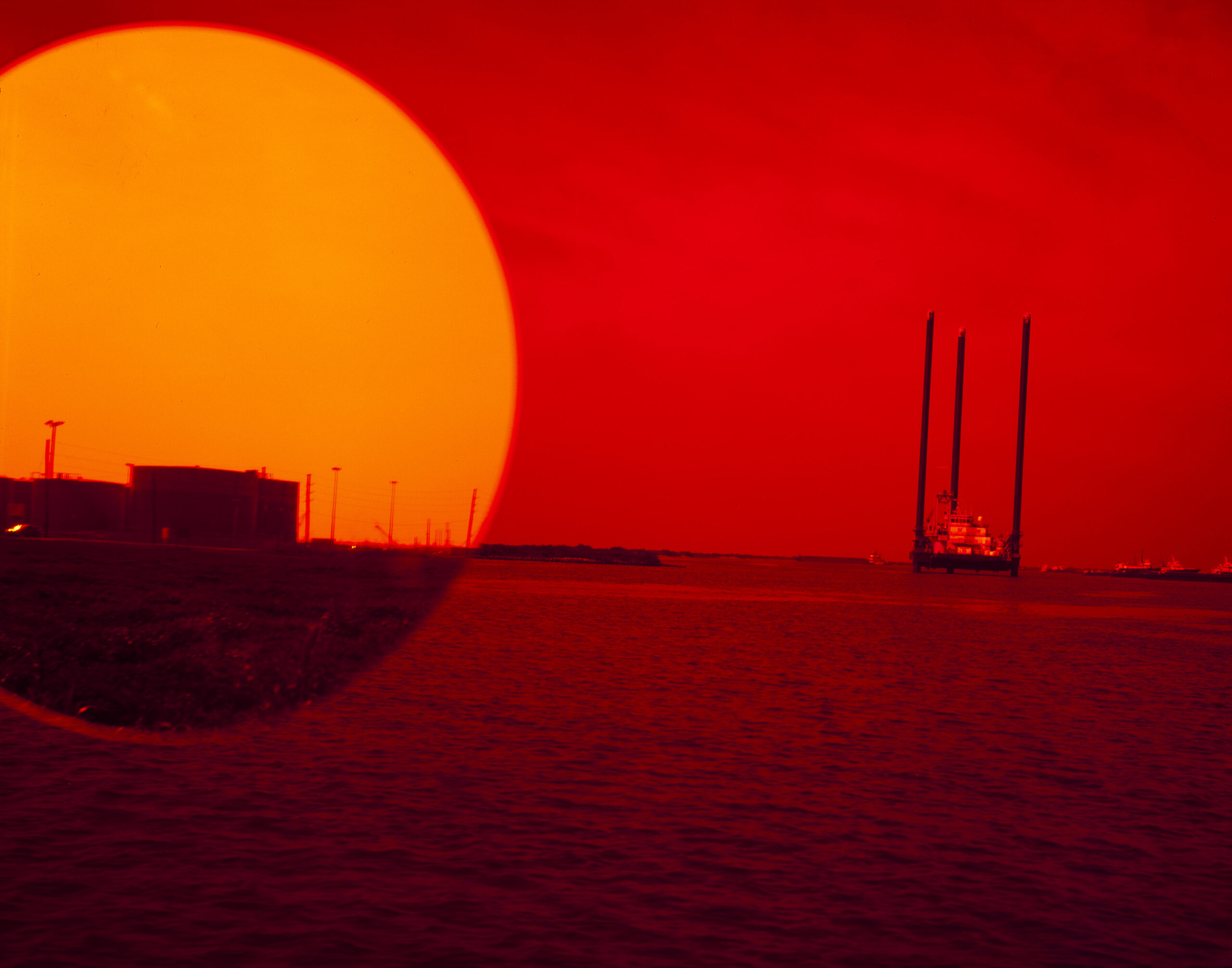 Photograph of a red sky and sea with boats at a distance. A large yellow circle on the top left resembles a sun setting over the ocean. Inside the circle, there is an image of a land mass with industrial buildings.