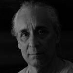 Black and white headshot of a man with pulled back grey hair and a white t-shirt.