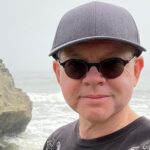 Headshot of a man wearing a grey ball cap, sunglasses, and a black shirt with white patterns. The beach and a large rock is in the background.
