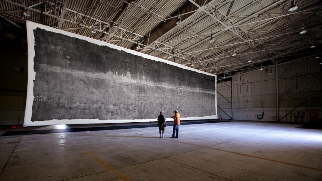 Two people standing in front of The Great Picture in a hangar.