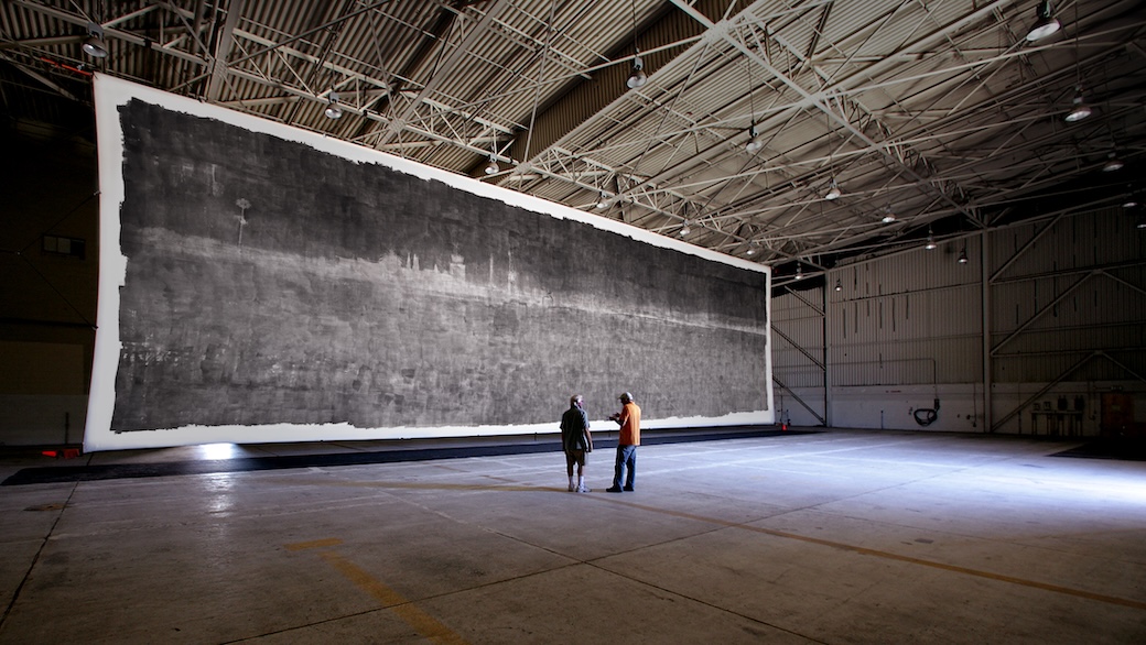 Two people standing in front of The Great Picture in a hangar.